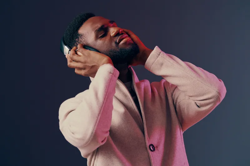 Young Black man enjoying music with headphones, expressing joy and relaxation in a stylish light beige coat against a grey background