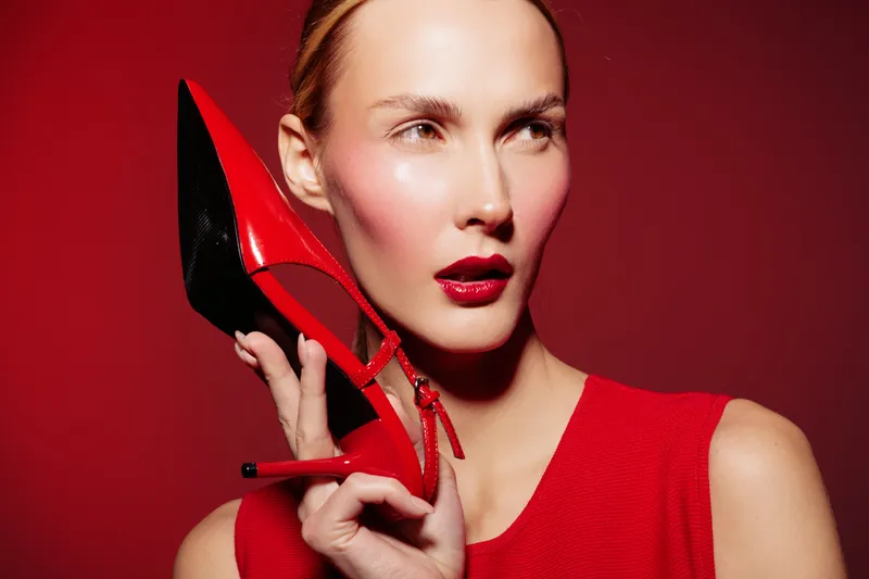 Woman in red dress holds red high heel shoe near face on red background. Fashion concept with bold makeup and stylish accessory for glamour and elegance.