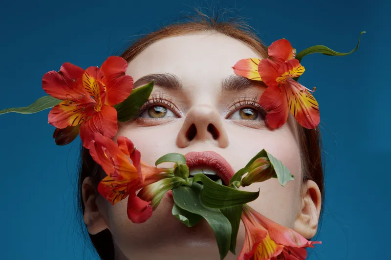 Woman with flowers on her face, expressing joy and tranquility against a blue background, symbolizing nature and beauty