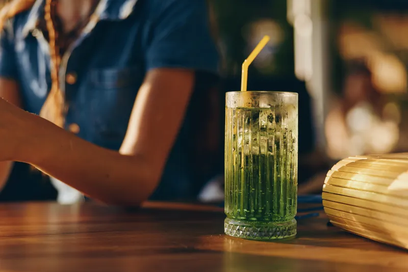 Modern boho-western denim street style woman enjoying a refreshing green cocktail in a film color warm indoor setting with lifestyle vibes.