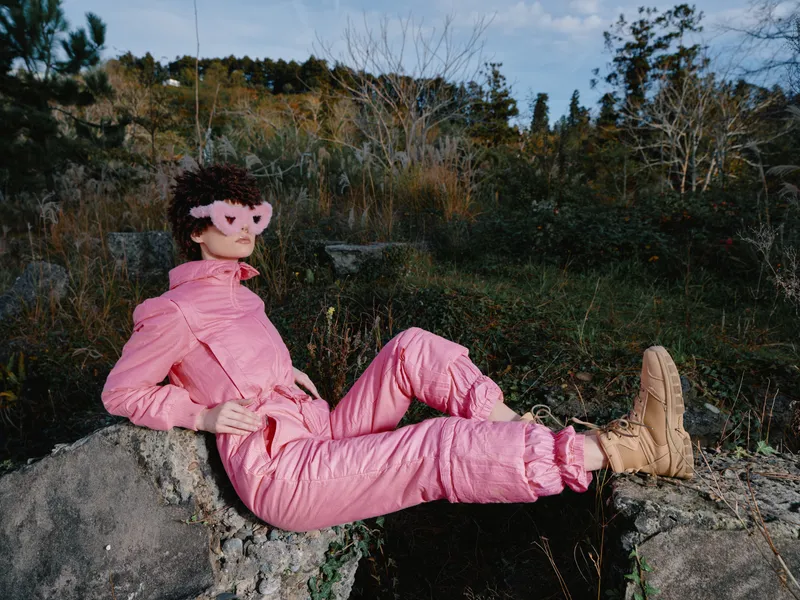 Pink suit cosplayer lounges on a rocky outcrop in an outdoor scene, wearing a pig mask and boots. The relaxed pose and vibrant color create a bold fashion statement amid rugged scenery.