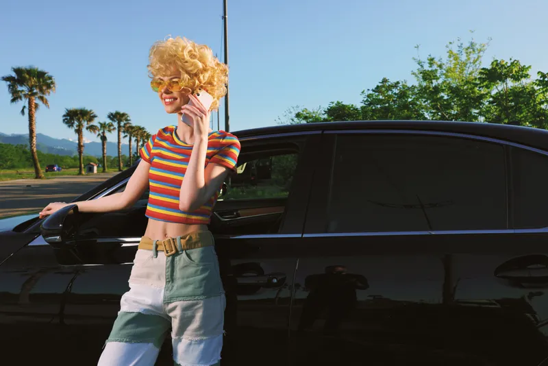 Young woman with curly blonde hair wears a striped crop top and light jeans, leaning against a sleek black car on a bright sunny day, exuding casual confidence and a carefree vibe.