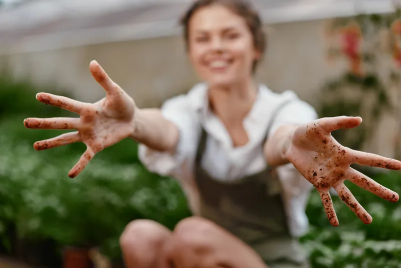 Young woman with dirty hands showcasing her love for gardening, wearing an apron in a vibrant greenhouse, expresses joy and connection with nature