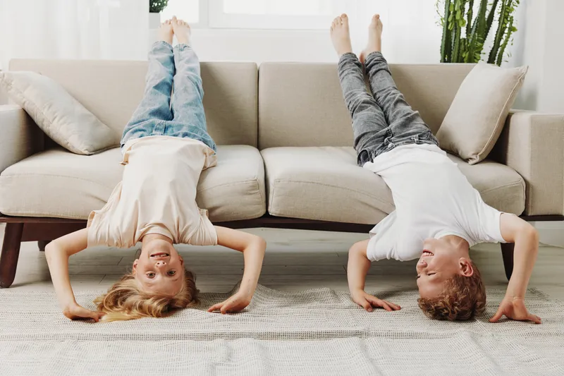 Two playful children in casual clothes do headstands on a carpet in a bright living room with beige sofa and plants. Concept of fun, childhood, and home activity.