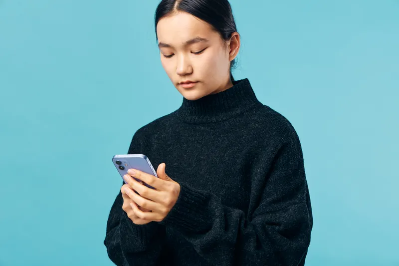 Young woman with dark hair wearing black sweater, looking down at smartphone with focused expression on light blue background in studio. Casual technology usage and communication concept.