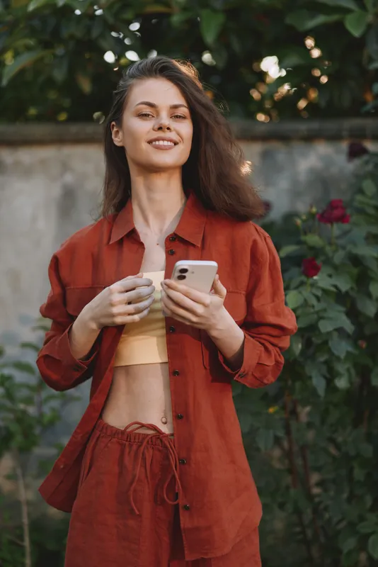 A young woman with curly hair wears a rust red matching set and smiles while checking her smartphone in a sunny garden
