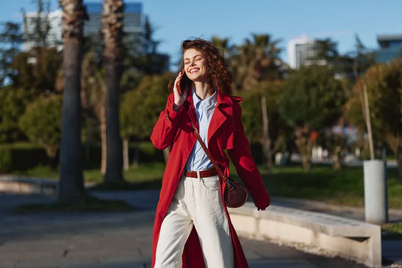 Young woman with curly hair wearing a red coat happily talks on the phone outdoors on a sunny day with palm trees in the background.