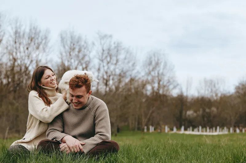 A couple sits on grass in a sunny park with their dog nearby, sharing a moment of affection and relaxation in the outdoors. A wholesome lifestyle scene that highlights companionship, nature, and