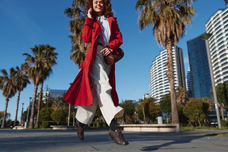 A young woman walks energetically down an urban street wearing a red coat, white wide-leg pants and brown boots under a bright blue sky and a bright, energetic vibe.