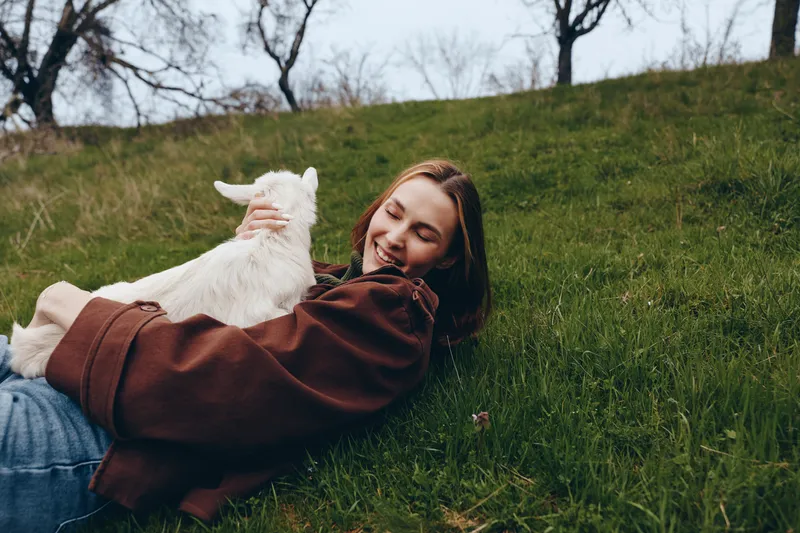 Cheerful woman outdoors, lying on grass, embracing a white goat kid, warm sunlight, relaxed pose, soft breeze, natural tones, lush greenery background, intimate countryside moment