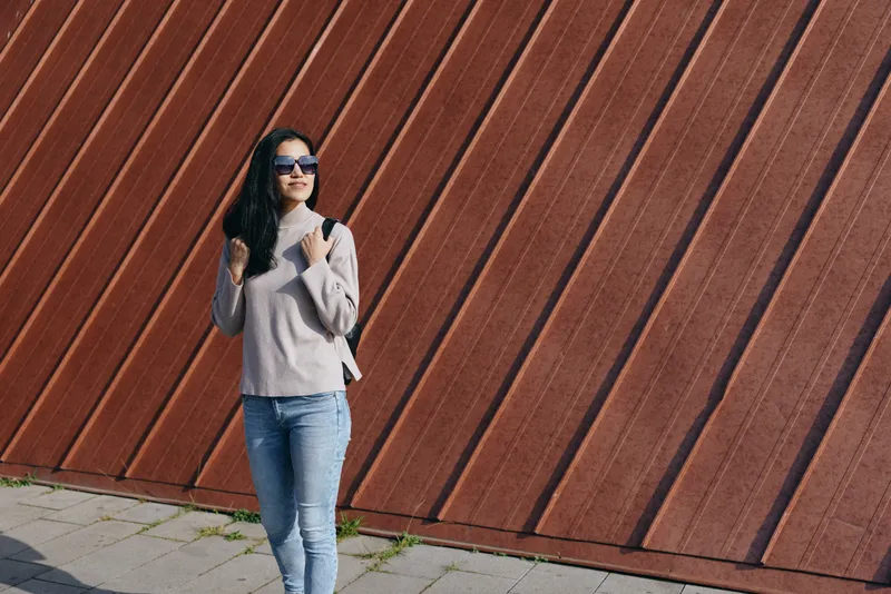 Woman wearing a cozy sweater and jeans, posing with sunglasses beside a diagonal wooden wall, urban outdoor fashion scene, confident relaxed style and effortless street chic