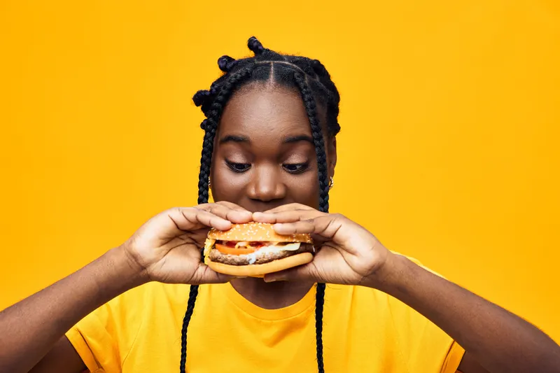 Young woman enjoying a burger with a colorful yellow background, showcasing excitement and vibrant lifestyle focused on healthy eating