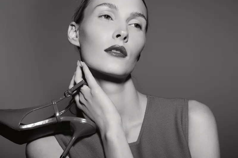 Portrait of a white woman with fair skin holding a high heel near her neck in a monochrome studio. Concept of fashion, elegance, and beauty.