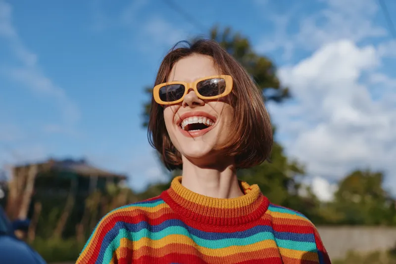 woman smiling striped sweater yellow sunglasses short hair outdoor sunny casual outfit happy lifestyle young adult laughing, wearing colorful knit, looking up in suburban park for travel and fashion