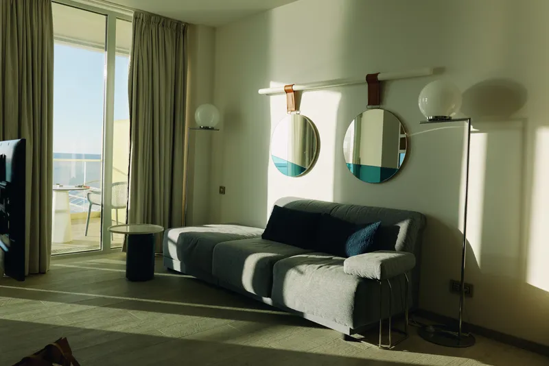 Modern living room with a sofa facing a balcony overlooking the sea, round mirrors, soft lighting and large windows creating a bright, inviting space for relaxation and contemplation.