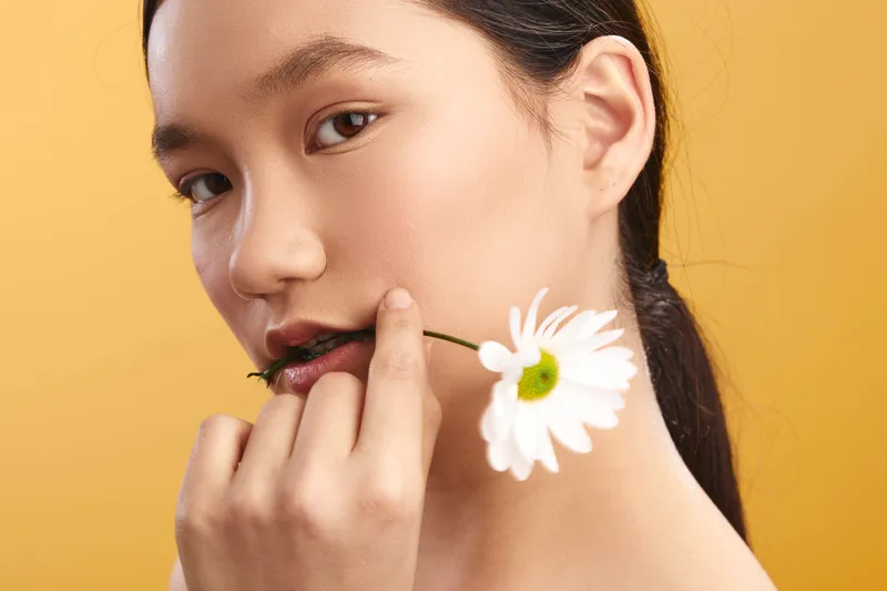 asian woman in a soft close up captures a delicate moment with a white daisy held near her lips against a warm yellow backdrop, conveying natural beauty and gentle expression.