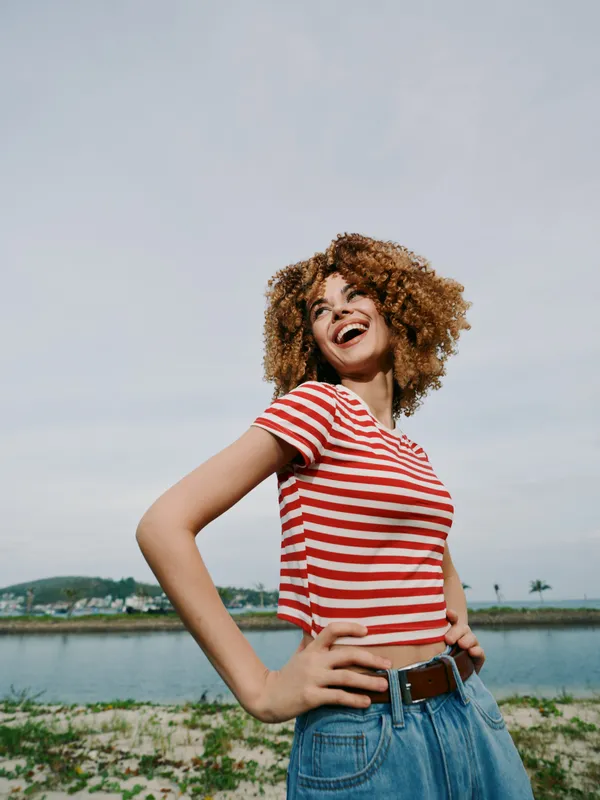 Young woman with curly hair stands outdoors near a waterway, wearing a red and white striped tee and denim jeans, exuding confidence, energy, and joyful charm.