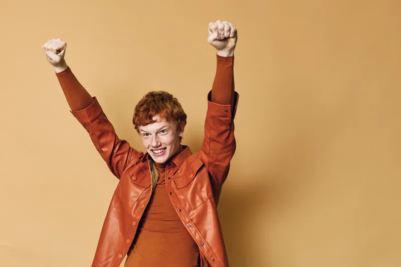 Happy young man with red hair wearing brown jacket and sweater celebrates success with raised arms and smiling face on beige background. Positive emotion and joy concept.