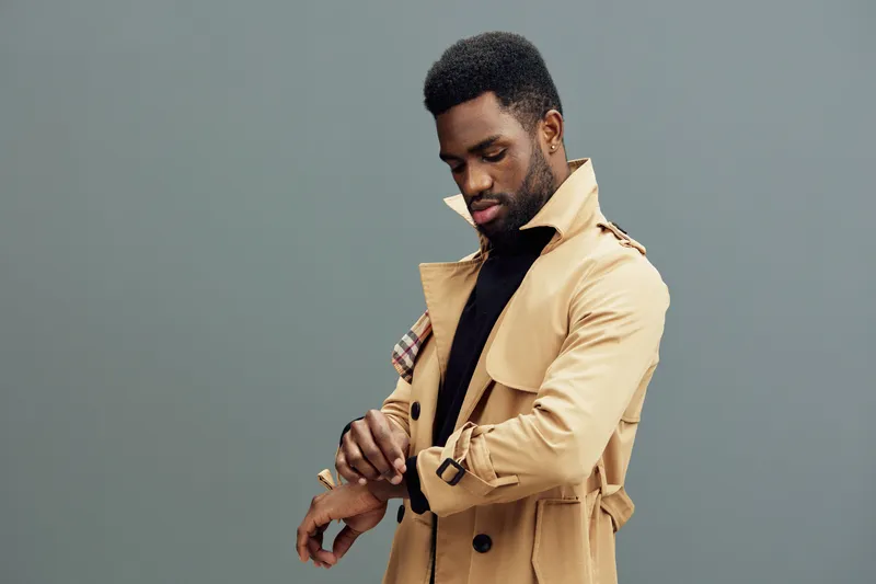 Stylish African man adjusting his trench coat against a minimalist grey background, conveying confidence and elegance, perfect for fashion and lifestyle themes