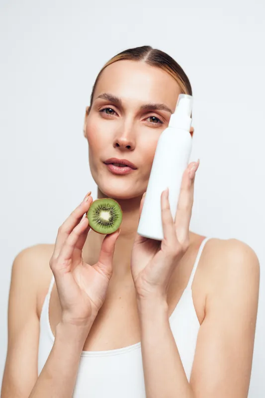 Young woman with long hair holds a white cosmetic bottle and a sliced kiwi close to her face while wearing a white tank top against a plain background.