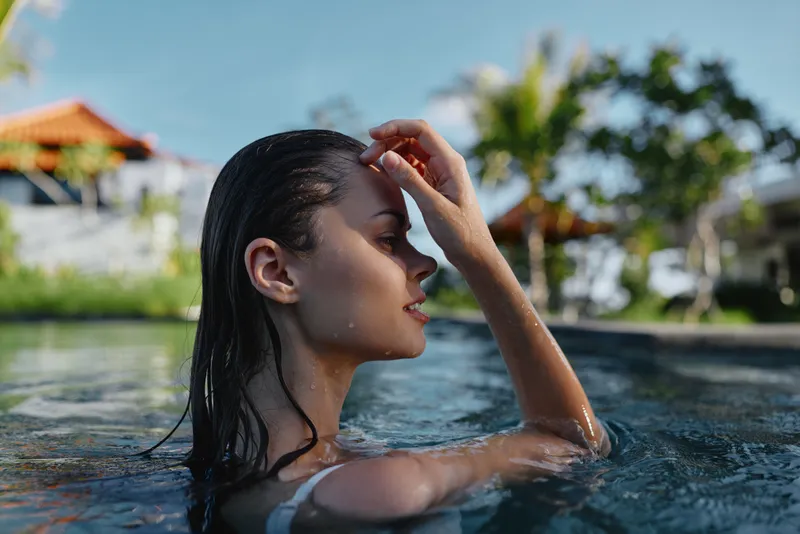 Relaxed woman in a pool, sunlit outdoor setting, profile view with wet hair, conveying summer leisure, wellness, and self care, ideal for lifestyle and beauty themes.