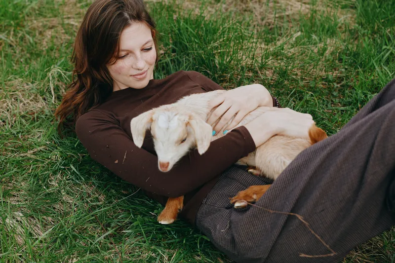 A relaxed woman lying on grass cradles a young goat kid in her arms, sunlit outdoors. A candid lifestyle moment showcasing bonding, care, and gentle affection in a peaceful rural setting.