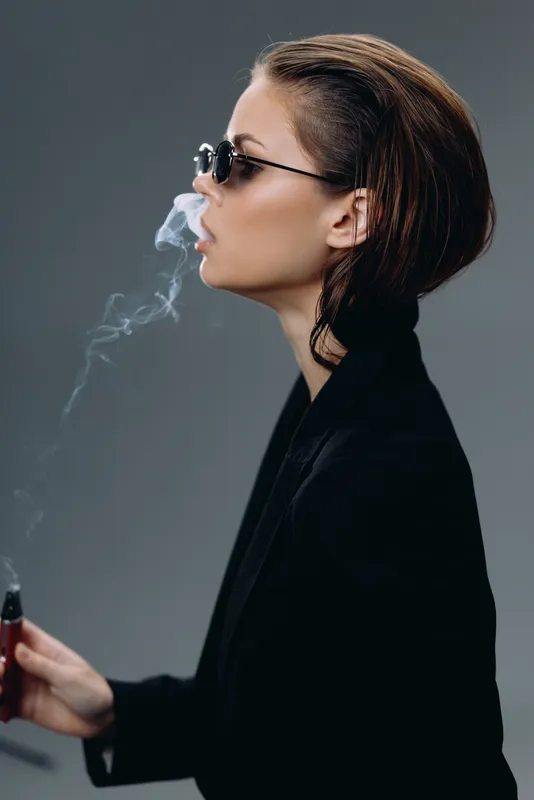 Stylish woman wearing sunglasses vaping electronic cigarette with calm expression and dark outfit