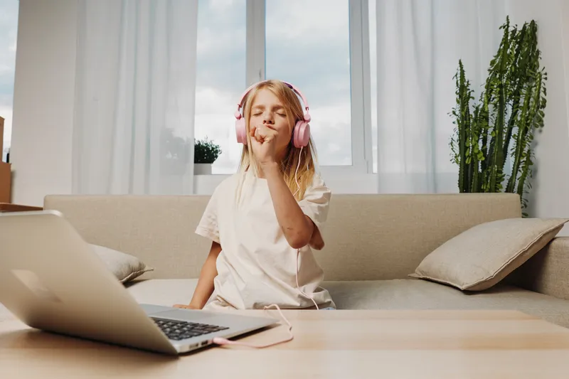 Young girl with headphones enjoys music with closed eyes sitting on sofa near laptop. Concept of relaxation, leisure, and digital entertainment at home.