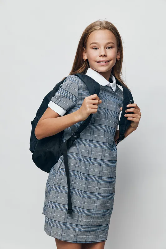 Girl child in school uniform with backpack standing and smiling on light background. Young student with long hair, education concept for back to school or learning time.