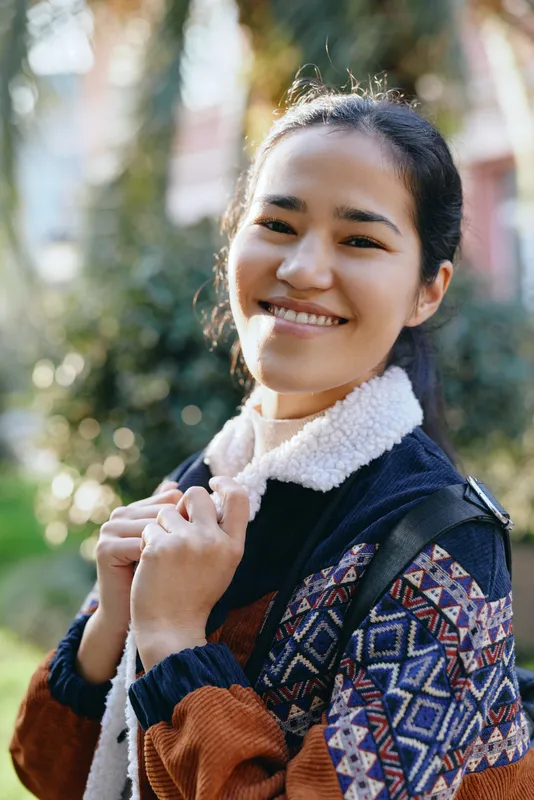 Smiling woman outdoors with backpack and patterned jacket, enjoying sunny weather. Casual fashion moment in a nature park setting, conveying friendly energy, travel readiness and relaxed urban style.