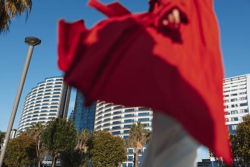 Young woman in a flowing red coat walks confidently in an urban setting with modern high-rise buildings and clear blue sky in the background.