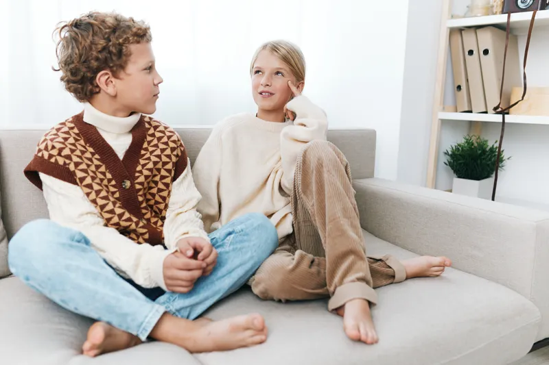 Two young adults sit on a light sofa wearing cozy knit sweaters, sharing a relaxed conversation in a bright living room with soft natural light and casual ambiance.