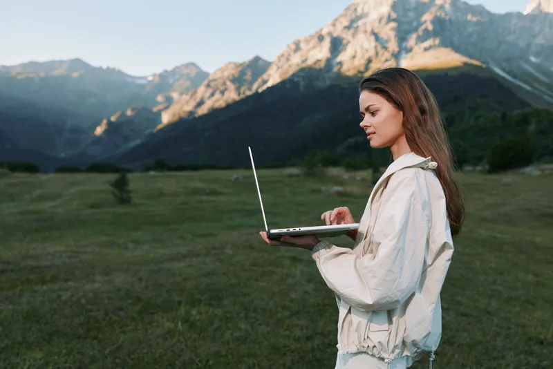 A focused outdoor professional uses a laptop in a scenic mountain meadow. She stands with calm concentration, blending technology with nature to create a serene and productive work setting.