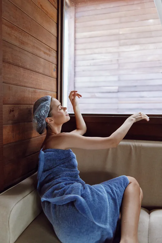 Relaxed woman wrapped in blue towel with shower cap sitting on beige sofa near wooden wall and window in cozy indoor setting, enjoying peaceful moment and calm atmosphere.
