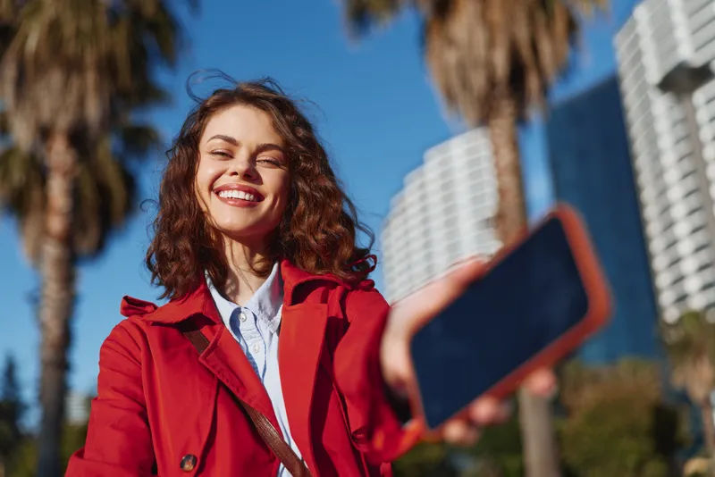 Smiling woman in a red coat takes an outdoor selfie in bright sunlight, urban cityscape behind her, casual fashion moment, cheerful mood, sunny day