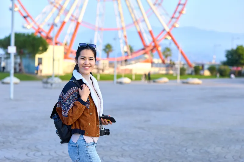 Woman outdoors with camera and backpack, smiling beside a colorful amusement park, showcasing casual fashion, travel vibe, and photography in a sunny day scene, urban exploration and joyful moment