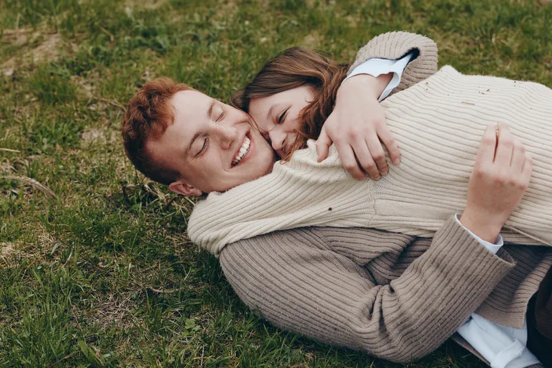 Two smiling people lie on grass, embracing warmly in casual sweaters, enjoying a candid outdoor moment of love, comfort, and carefree lifestyle in a sunlit park, sharing laughter, closeness, and a