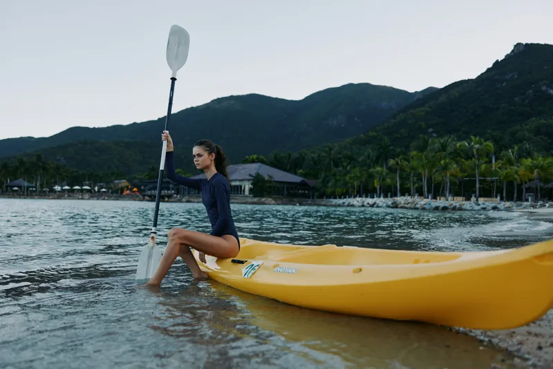 Young woman kayaking in calm water near tropical shore with mountains during early morning, showcasing outdoor adventure and active lifestyle.