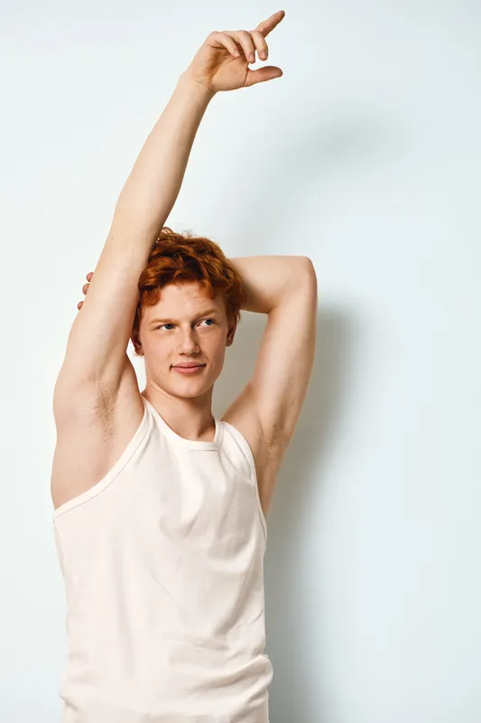 Young man with red hair wearing white sleeveless shirt stretching arm upwards and looking to side on white background. Casual pose with natural expression.
