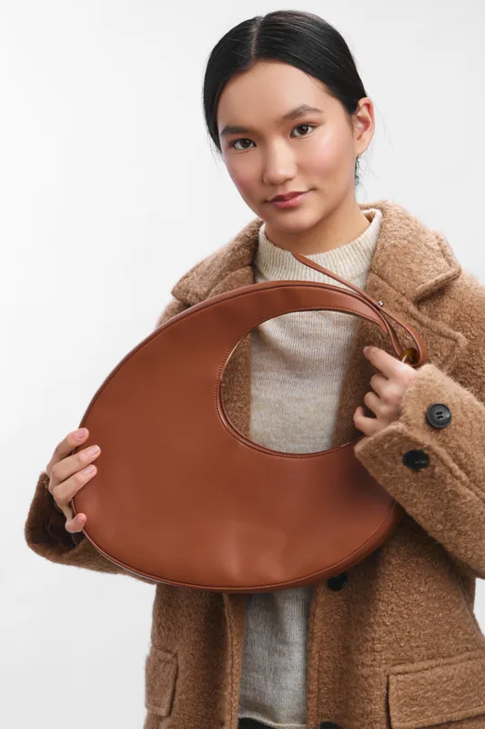 asian woman wearing a warm beige sweater and a stylish coat holds a brown circular handbag, posing confidently in a clean studio setting. soft light highlights texture and fashion detail.
