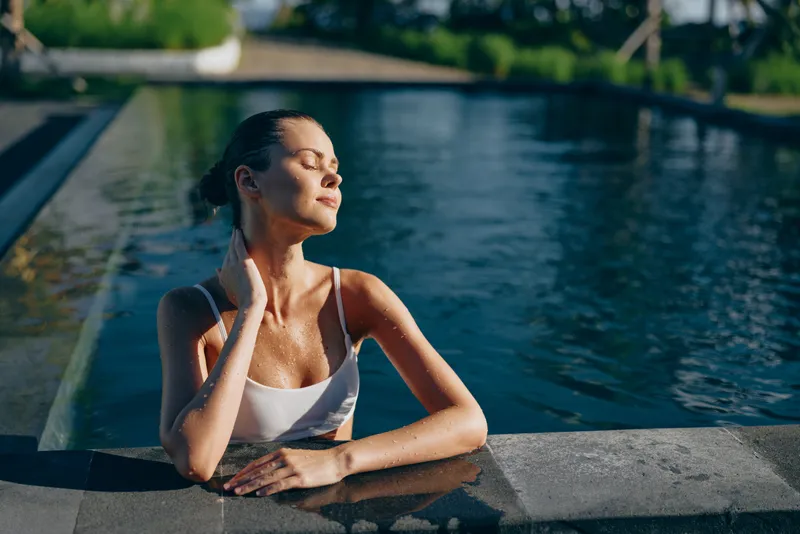 Relaxed woman in a white swimsuit rests at pool edge, sunlit moment, calm expression, soft water reflections, warm breeze, casual lifestyle, lush outdoor backdrop and clear blue sky above today