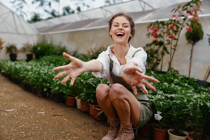 Joyful female gardener in a greenhouse, wearing casual clothing, surrounded by vibrant green plants, showcasing happiness and connection with nature