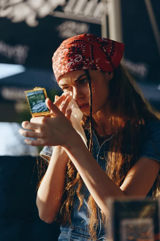 young woman with red bandana applying makeup using compact mirror outdoors in natural sunlight during daytime