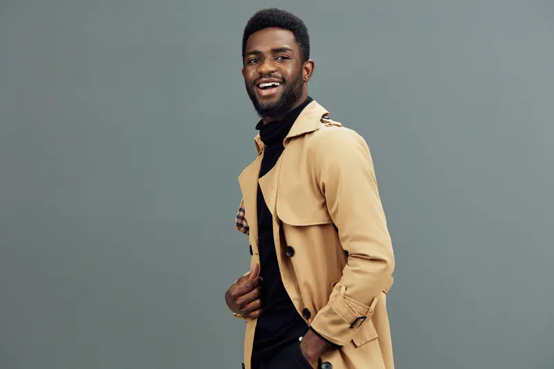 Stylish young Black man smiling in a beige trench coat and black turtleneck against a grey background, exuding confidence and fashion sense