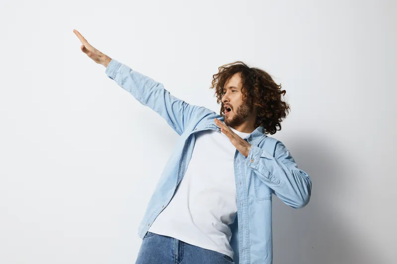 Young man with curly hair in a light blue shirt showcasing an expressive dance move against a neutral background, conveying joy and energy in a stylish manner