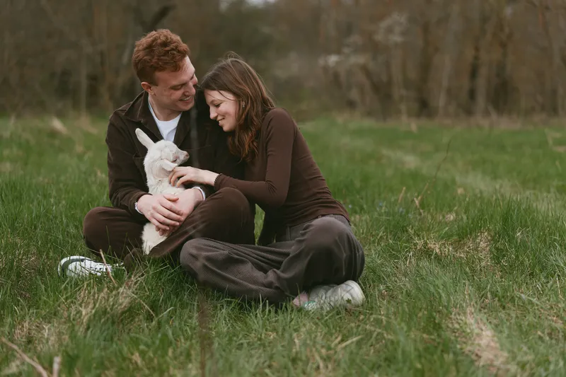 A smiling couple sits on grass outdoors, petting a small white dog, enjoying a peaceful moment together in a sunny park, capturing everyday affection and relaxed lifestyle
