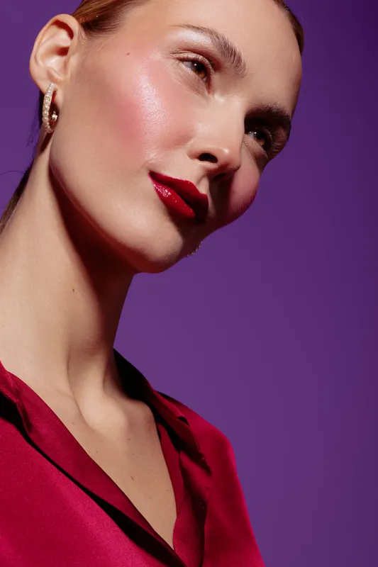 Close-up of a woman with glowing skin and red lipstick wearing a red blouse against a purple background. Concept of beauty, makeup, skincare, and fashion portrait.