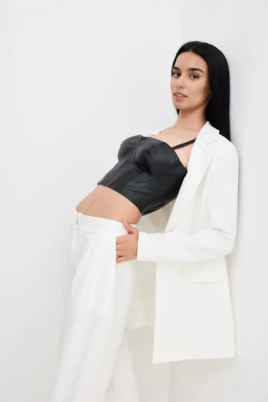 stylish woman wearing a black leather corset and white blazer poses confidently against a plain white background in a modern fashion studio setting