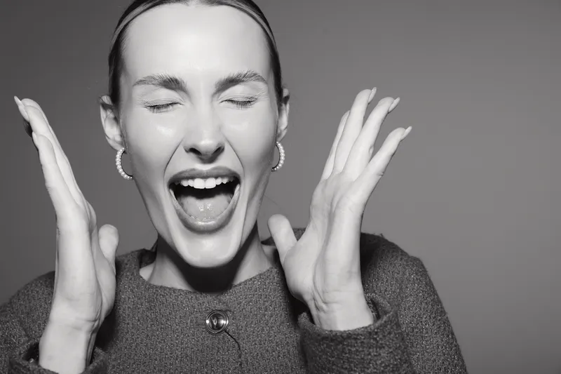 A young white woman with light skin and closed eyes expresses excitement with open mouth and raised hands in black and white photo.