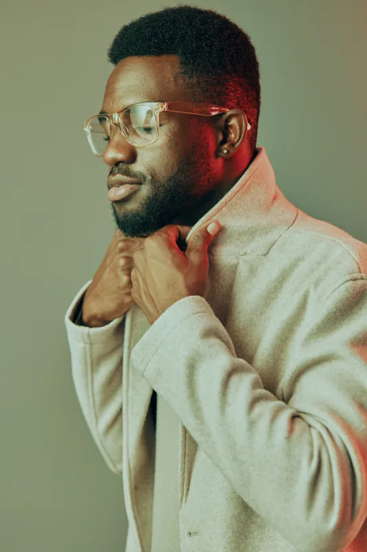 Stylish young man in a beige coat, wearing glasses, conveys confidence and elegance against a soft, muted background with warm hues